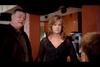 A man and a woman are standing indoors, facing another person whose back is turned to the camera. The woman is wearing a black top, while the man is dressed in a dark shirt and jacket. The setting appears to be a restaurant or a cafe.