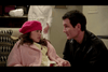 A young girl wearing a red beret and a pink coat sits, looking at a man in a black jacket who kneels beside her, smiling. Another person in a white outfit stands in the background. The setting appears to be indoors, possibly a home.
