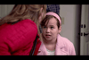 A young girl with a headband looks up while speaking to a woman with light brown hair who is partly facing away. The girl is wearing a light pink jacket, and the woman has on a bright pink jacket. They are indoors with a blurred background.