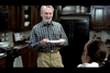 An elderly man with white hair and a white beard wears a checkered shirt and khaki pants while standing in a kitchen, holding a plate. He is smiling and looking at a young person with long hair who has their back to the camera.