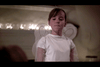 A young child dressed in a white outfit stands indoors, with a background of soft lighting that appears to be festive or decorative. The child has their hair tied back and seems to be looking forward. The setting suggests a special occasion or gathering.