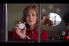 A woman with light brown hair, wearing a red top, holds up a small circular white object while looking out of a window. The background is decorated with Christmas ornaments and garlands, including a wreath.