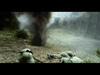 A dramatic scene with soldiers in army uniforms taking cover on the ground as a large explosion sends debris into the air in a forested area. Smoke and dust obscure the background, adding to the intensity of the moment.