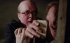 A man with glasses holds another man's head against a wooden surface, pressing a piece of paper tightly against the second man's face. The scene appears intense and dramatic, suggesting a moment of confrontation or interrogation.