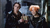 Two people with spiky, colorful hair and leather jackets are standing in an industrial setting. One of them is holding a small, intricate box with both hands, while both are smiling. The background shows large pipes and metal structures.