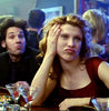 A woman with long, wavy hair leans on her hand with a bored expression at a bar counter, wearing a red dress. A man beside her makes a playful face, puffing his cheeks out. The background shows other bar patrons and a blue-lit environment.