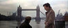 Two people are conversing by a river with the iconic Tower Bridge in London in the background. One person is seated, and the other is standing, wearing a light-colored coat. The sky is overcast, giving a moody atmosphere to the scene.