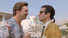 Two men are having a face-to-face conversation outdoors. The man on the left is wearing a floral shirt and has light-colored hair, while the man on the right is wearing a yellow jacket, sunglasses, and has dark hair tied back. A building and trees are in the background.