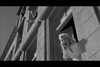 A woman with wavy hair is leaning and shouting out of a window of an urban building. The scene is in black and white, capturing dramatic expressions and architectural details of the structure.