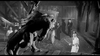 A black-and-white scene featuring multiple characters in a dramatic confrontation. A large, imposing, dragon-like creature stands in the foreground facing a group of people holding weapons. The surroundings resemble an old wooden structure. The mood is tense and intense.