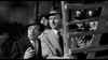 Two men in suits and hats look surprised, standing near a wooden ladder. In the background, a group of similarly dressed individuals are also visible. The image is in black and white and appears to be from a vintage film or scene.
