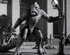 A black-and-white image shows a large stop-motion animated dinosaur-like creature terrorizing a city street. Tiny model buildings, cars, and people are seen in the background, adding to the scene's cinematic drama.