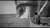 A black and white scene shows a person in a small boat rowing away from a gigantic creature with sharp teeth emerging from the water. The creature's mouth and part of its body are visible, creating a dramatic and intense atmosphere.