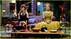 Two women are sitting on a couch in a colorful, eclectic living room. The woman on the left is wearing a black dress, and the woman on the right is dressed in a yellow outfit with a headpiece. A coffee table with various items is in front of them.