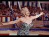 A woman with blonde hair is standing in front of a circus-like setting with a blue, red, and white striped backdrop. She is wearing a strapless zebra print dress and has her arms spread wide open, appearing elated. An audience is visible in the background.