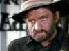 A close-up of a man with a serious expression, rugged beard, and dark hair wearing a weathered hat and dark clothing. The background is slightly out of focus, with indistinguishable objects.