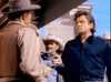 Two men in a western setting face each other. The man on the left wears a cowboy hat, a brown vest, and a tan shirt. The man on the right, dressed in a dark blue shirt, is looking intently at the other. A person in a lighter cowboy hat stands in the background.
