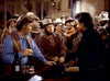 A group of people, mainly men, dressed in old Western attire are gathered in a saloon. The focal point is on two men facing each other, engaging in a conversation. The scene includes rustic wooden furniture, bottles, and a dimly lit interior.