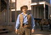 A person stands in a western-style town dressed in old-fashioned cowboy attire, including a brown vest, blue shirt, and cowboy hat. The background features wooden buildings and a horse. The sun casts shadows, highlighting a rustic, historical setting.