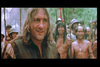 A man with shoulder-length hair is smiling in the foreground. He is wearing a dark tunic and is surrounded by indigenous people with darker skin and traditional attire. The background features trees and a group of people, possibly in a forest or jungle.