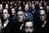 A group of serious-looking individuals are seated closely together, all staring intently in the same direction. They are dressed uniformly in dark clothing, and their expressions appear focused or apprehensive. The scene is dimly lit, casting shadows on their faces.