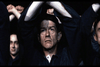 A man with a serious expression stands with his arms raised overhead, interlocking his hands with others in a crowded, shadowy environment. The lighting casts dramatic shadows on their faces, creating a tense and somber atmosphere.