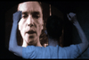 A person is seen from behind, watching a large screen displaying another person's face. The screen shows a close-up of the face, while the viewer's raised arms are partially visible. The overall scene is dark, with the screen being the primary light source.