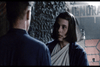 A woman with shoulder-length hair, wearing a white wrap and blue robe, stands in front of a man whose back is turned. They appear to be in a dimly lit, industrial setting with rough-textured walls and a partially visible warning sign in the background.