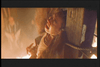 A person dressed in historical clothing appears to be in distress, lying on the ground with their upper body propped up against wooden beams. The scene is shrouded in dim lighting and surrounded by a smoky or fiery atmosphere.