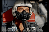 A person wearing a full pilot's helmet and oxygen mask is seated in what appears to be a cockpit. The individual is dressed in a flight suit, with various patches and equipment straps visible. The background includes the inner walls of the aircraft.