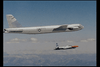 A military jet with a gray fuselage and distinctive circular insignia on the tail flies above a mountainous landscape. Below and slightly behind, a smaller aircraft with an orange nose cone also flies. Both planes are set against a blue sky with scattered clouds.
