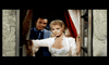 A woman in a white dress with a plunging neckline stands by an open window, holding the frame with one hand. A man in a blue suit stands behind her, partially obscured by a red curtain. They are in an ornate, possibly vintage, setting.