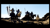 A group of four riders on horseback wielding spears and other weapons advances across a rocky terrain with bright blue sky in the background, creating a dramatic silhouette effect.
