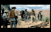 A group of cowboys stands in a desert landscape with rugged terrain under a partly cloudy sky. They are dressed in traditional western attire, including hats and boots, and appear to be engaged in conversation. Horses are present, and a person lies on the ground nearby.