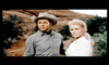 A man and woman, both dressed in western attire, stand outdoors with a rocky landscape and foliage in the background. The man wears a cowboy hat and looks off into the distance, while the woman, with blonde hair, looks slightly to the side.