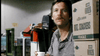 A person with a mustache and casual shirt holds a cup while sitting near stacked boxes labeled "COFFEE HOUSE BLEND." The background shows a partially visible office or workplace setting with various objects and equipment.