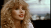 A person with long, curly blonde hair and bright red lipstick is gazing to the side. The background is blurry, hinting at an outdoor location with blue and green hues. The individual appears to be engaged in conversation or deep thought.