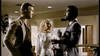 Three people are engaged in conversation in what appears to be an indoor setting. One man on the left is wearing a beige jacket, a woman in the middle is wearing a white dress, and another man on the right is wearing a dark suit. The background is blurry.