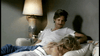 A person with a mustache, wearing a white t-shirt, sits on a couch and looks at another person lying down with their back toward the camera. A lamp is on a small table next to the couch, illuminating the scene in a dimly lit room.