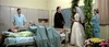 A hospital room features a patient lying in bed with medical equipment nearby. At the foot of the bed, a bride and groom stand with a priest, seemingly conducting a wedding ceremony. Two medical staff members are present, one near the bed and another near the ceremony.