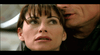 A woman with short, dark hair looks upward with a concerned expression, while a man close behind her is speaking softly near her ear. Both are wearing dark clothing, and the background is blurred.