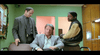 Three men are in a small office with green walls. One man is seated in a chair in the center, wearing a gray suit. The man on the left is standing and looking down at him, wearing a dark suit, while the man on the right leans against a desk, wearing a beige and black jacket.