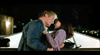 A man and a woman are sharing an affectionate moment outdoors at night. The man, wearing a denim jacket, leans in towards the woman, who is dressed in a purple sweater, as she gazes up at him. The background features soft lighting and blurred greenery.