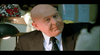 A bald man in a suit and tie is sitting at a desk, looking attentively at someone off-camera. The background includes a desk lamp and some papers pinned to a bulletin board.