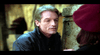 A man with short, light brown hair and wearing a dark coat is leaning close to another person who is wearing a maroon beret. They are outdoors with a blurred background featuring stone structures. The scene suggests an intimate conversation.