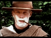 A person wearing an old-fashioned wide-brimmed hat and a tan shawl or coat is standing outdoors against a green leafy background. The person has a white beard and is smoking a pipe, exuding a vintage, rustic appearance.