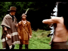 Three men are depicted in an outdoor setting. Two of them stand facing each other; one wears a brown poncho and hat, the other a brown fringed outfit with a headband. The third man, with his back to the camera, is shirtless and appears to be holding a bow.