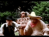 Three people are outdoors in a wooded area. One holds a "DETOUR" sign, dressed in an unbuttoned vest and hat. Another person, wearing a large sombrero and brown outfit, appears to be sitting in a makeshift cart. The third person is blurred and indistinct.