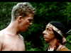 A scene featuring two individuals standing close to each other in a forested area. One person on the left is shirtless with short blonde hair, looking intently at the other person who is wearing a headband with feathers and traditional attire, depicting a Native American.