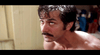 A shirtless man with wet hair and a mustache looks upward with a concerned expression. The background appears to be a bathroom with blurred shelves and a white door. The lighting is soft, giving the scene a dramatic and tense atmosphere.